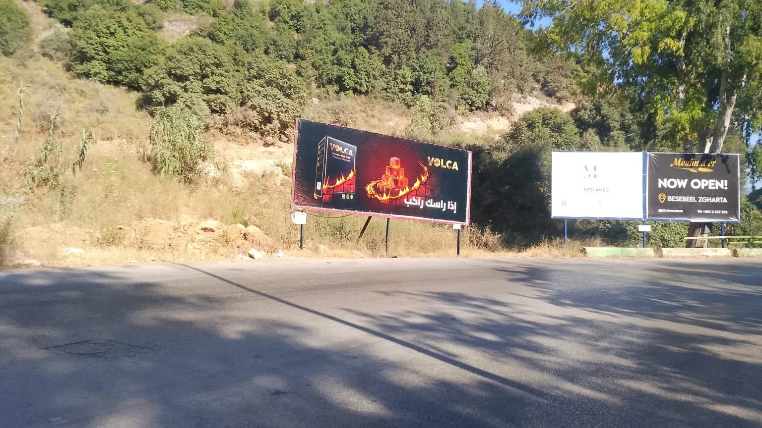 Volca landscape billboard on a hillside road beside other boards, إذا راسك راكب